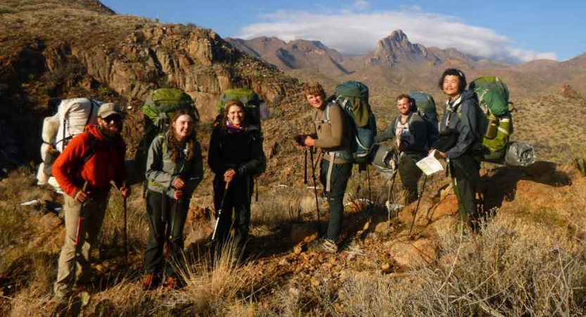a group of students carrying backpacks stand amidst a desert landscape with mountains in the background